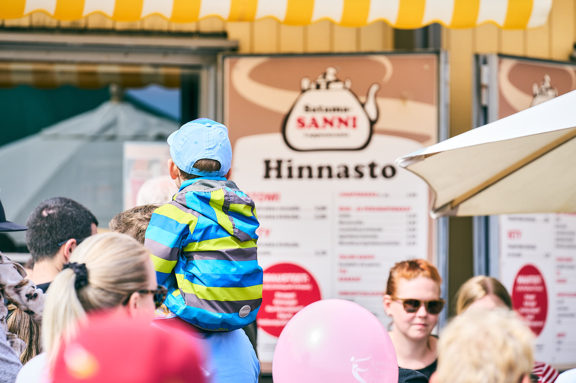 Ihmisiä keltaisen vetykojun edessä. Jonottamassa aikuinen, jonka hartioilla istuu lapsi, jolla on raidallinen takki.